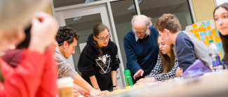 A professor is standing at a table surrounded by undergraduate students in a discussion.