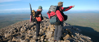 Two people standing atop a rocky mountain wearing outdoor gear. One man is pointing into the distance while a woman stands beside him.