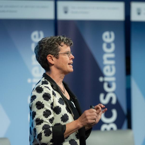 A professor standing in front of a row of UBC Science banners speaks to an audience off screen.